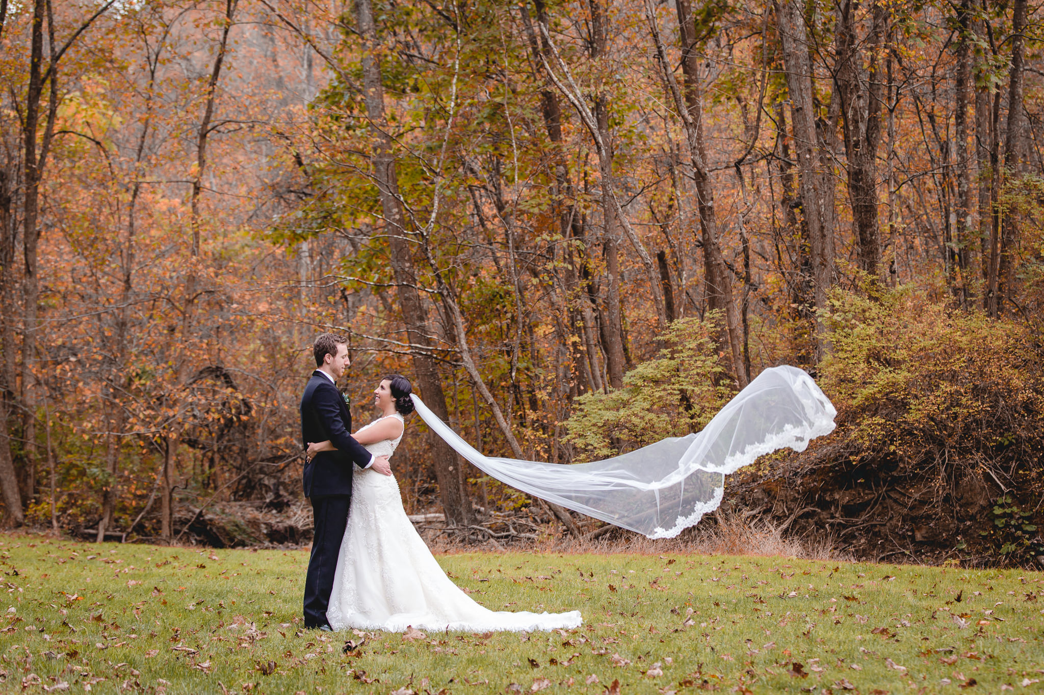 Bride and groom stand in the park with her cathedral veil blowing in the wind
