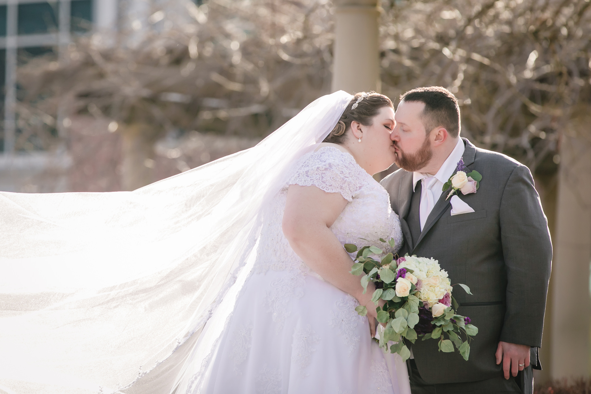 Bride's cathedral veil blows in the wind as she kisses her groom at Robert Morris University
