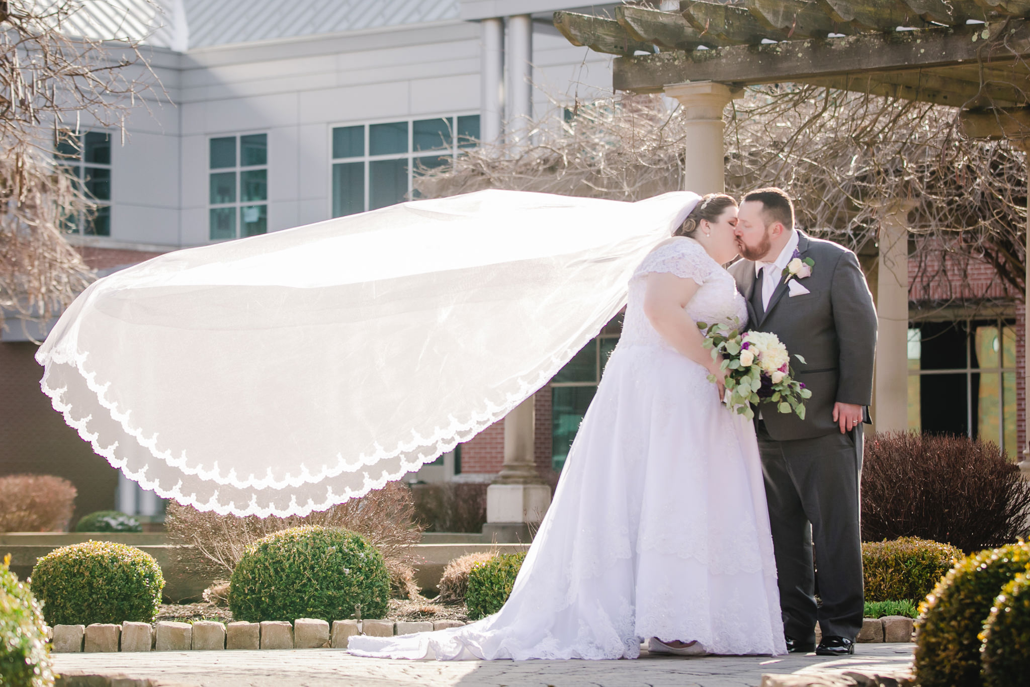 Bride's cathedral veil floats behind her as she kisses her groom at Robert Morris University