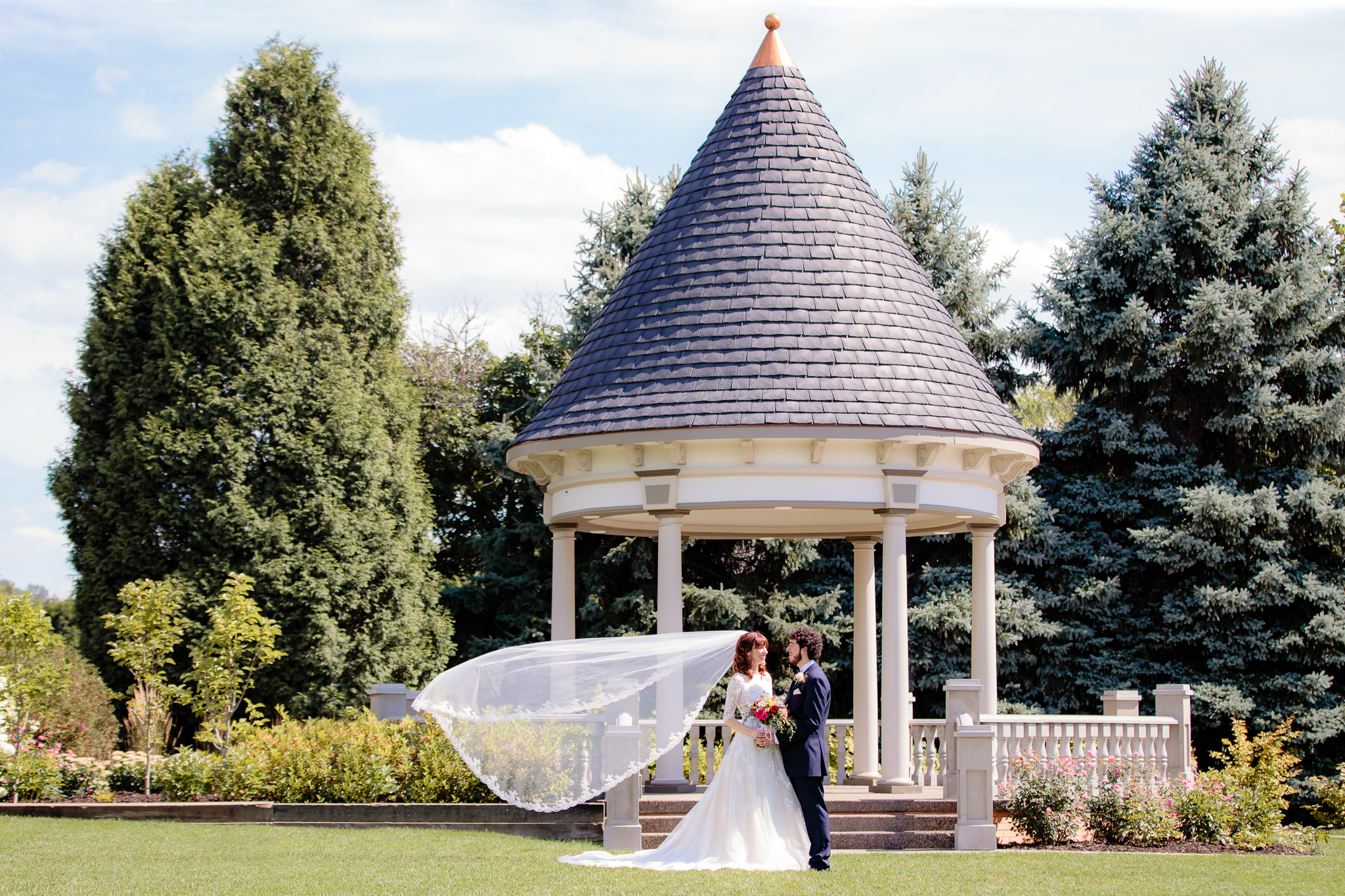 Bride's cathedral veil blows in the wind in front of the gazebo at Beaver Station