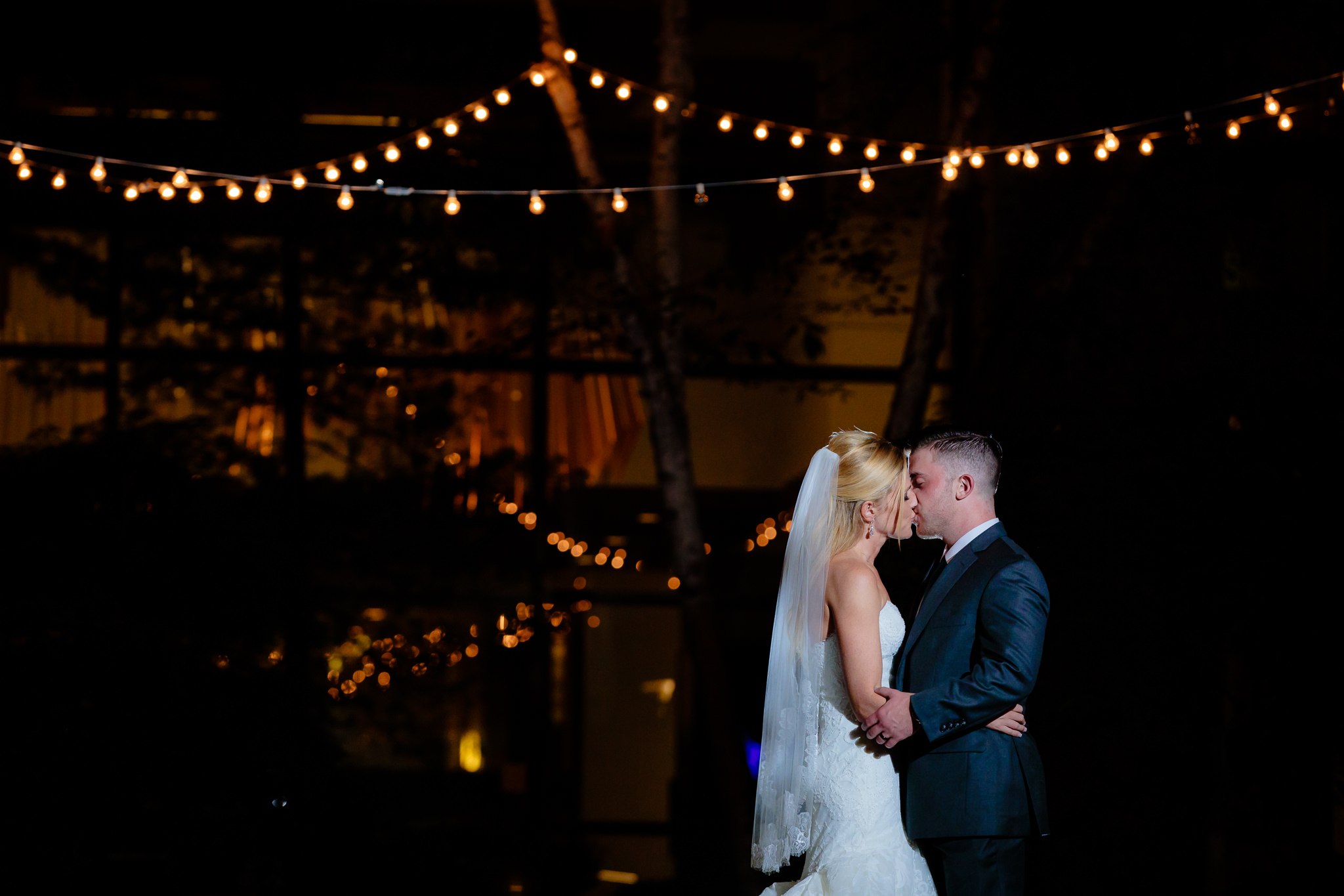 Bride and groom kiss under string lights at night in the Pittsburgh Airport Marriott courtyard