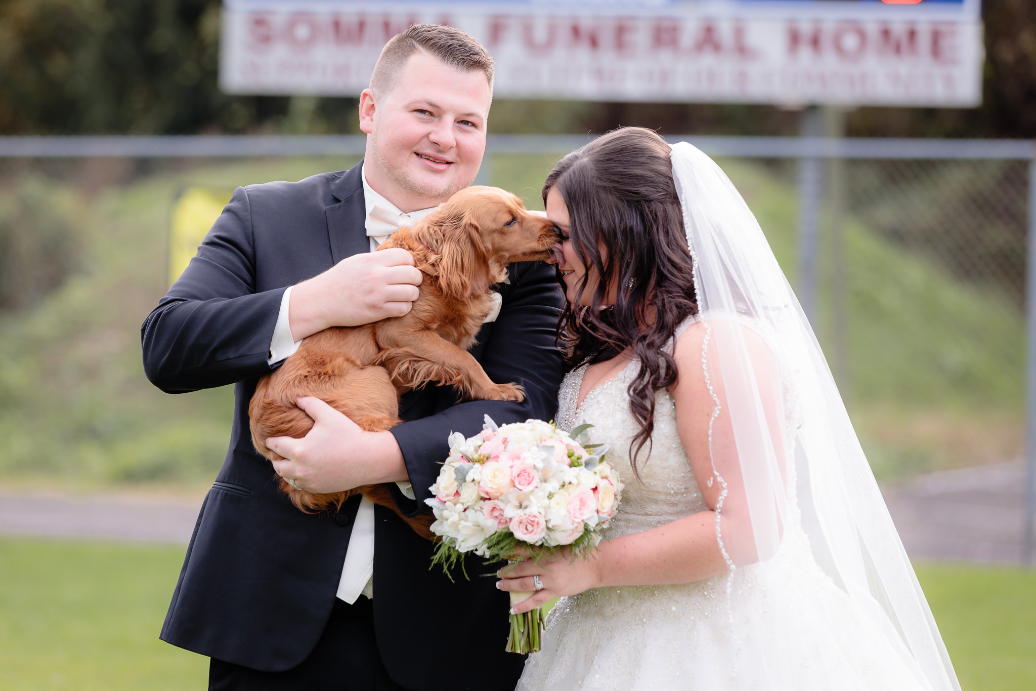 Bride's dog licks her face during portraits at the Robinson Township Girls Softball field