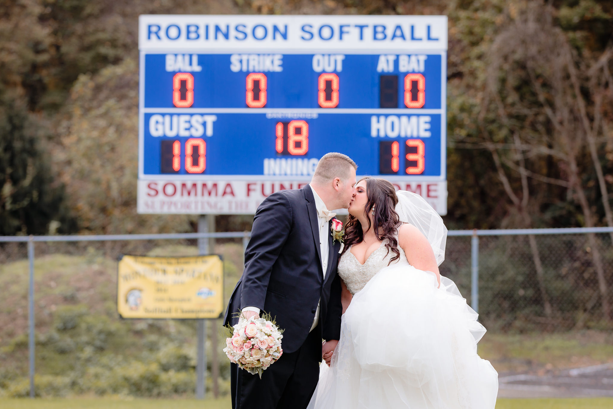 Bride & groom kiss in front of the scoreboard at the Robinson Township Girls Softball field