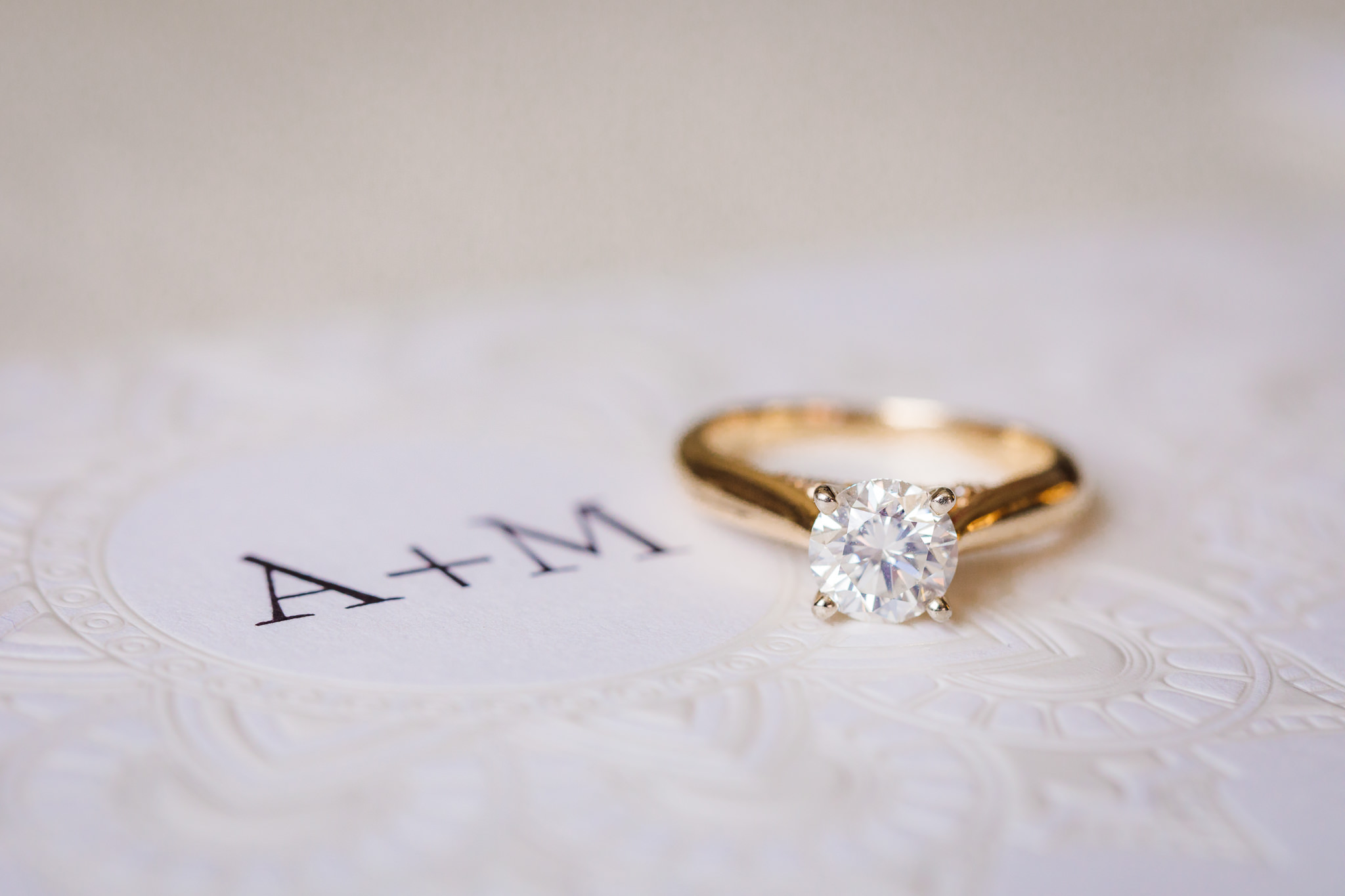 Gold diamond engagement ring rests on the monogram of a custom white wedding invitation