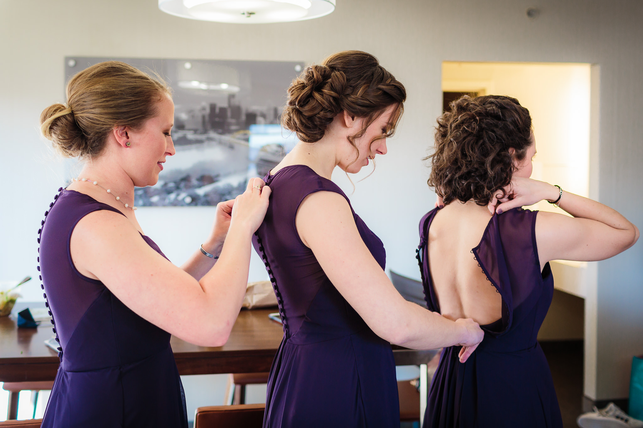 Three bridesmaids button the back of each other's purple Bill Levkoff bridesmaids dresses