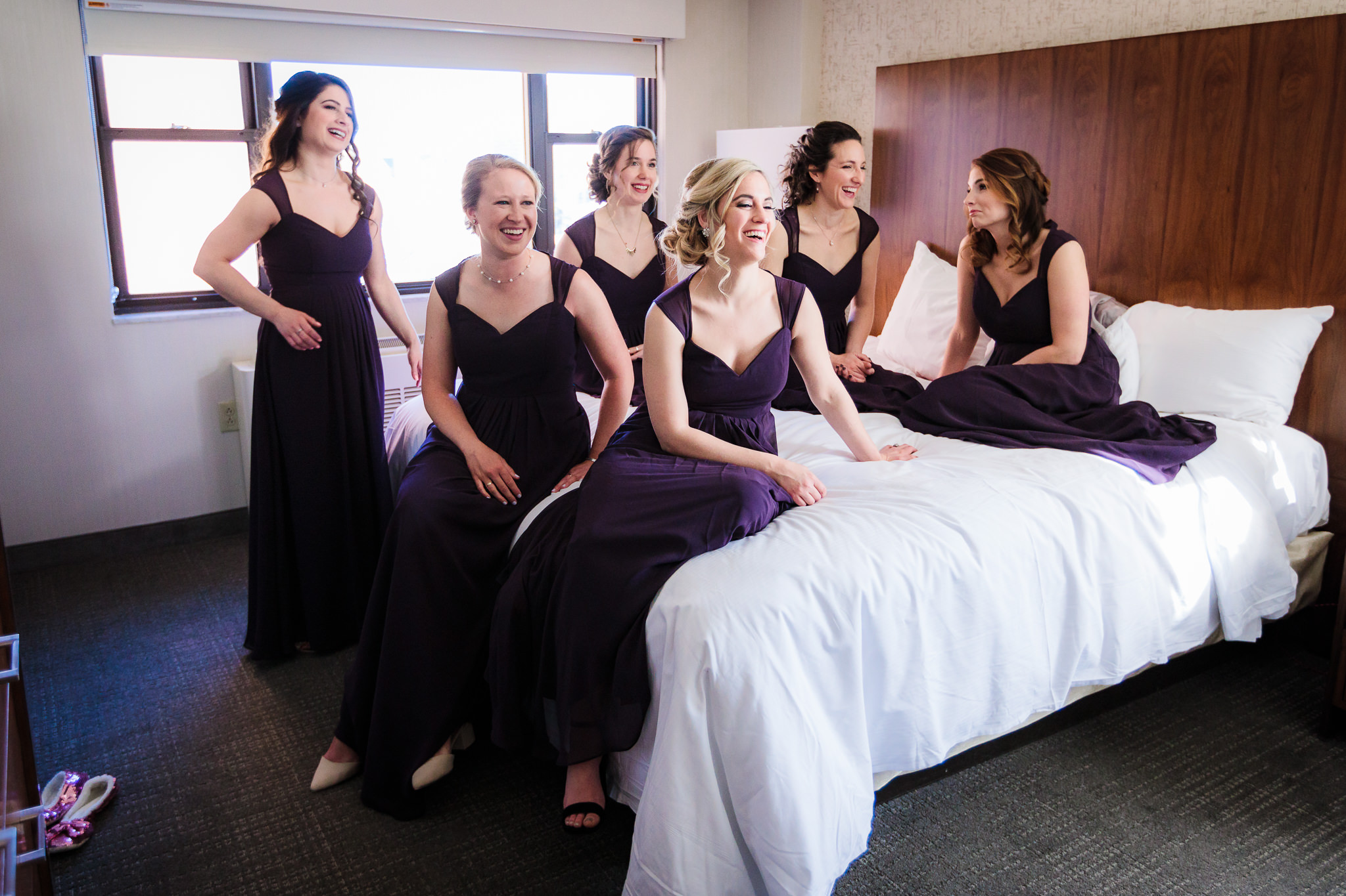 Bridesmaids in purple Bill Levkoff dresses watch as bride gets into her wedding dress