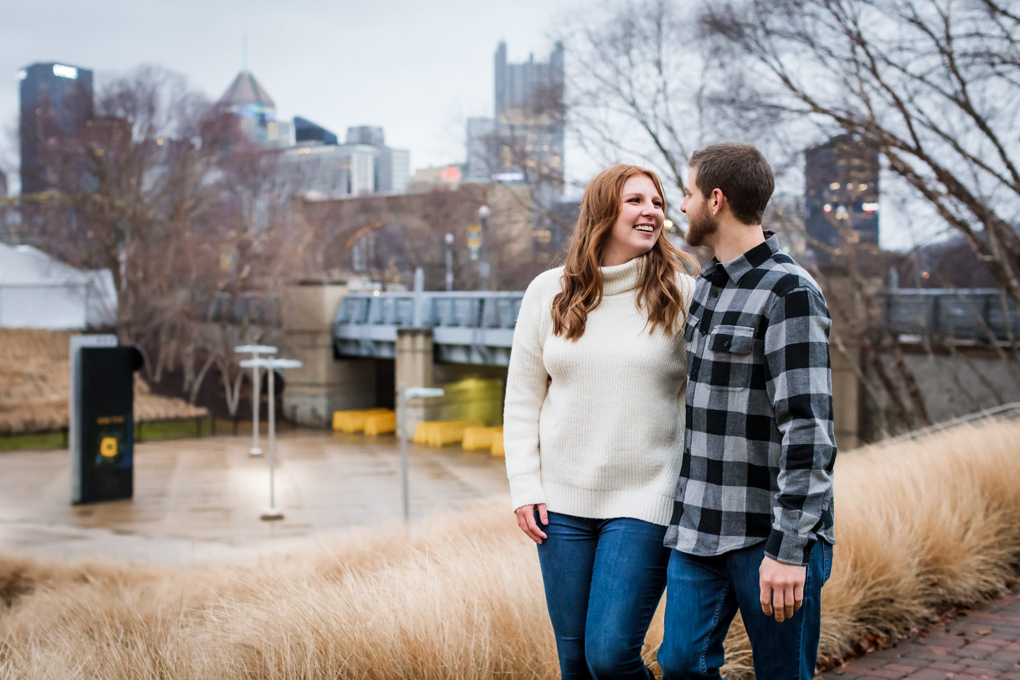 Couple walks with Pittsburgh skyline behind them at a North Shore engagement session